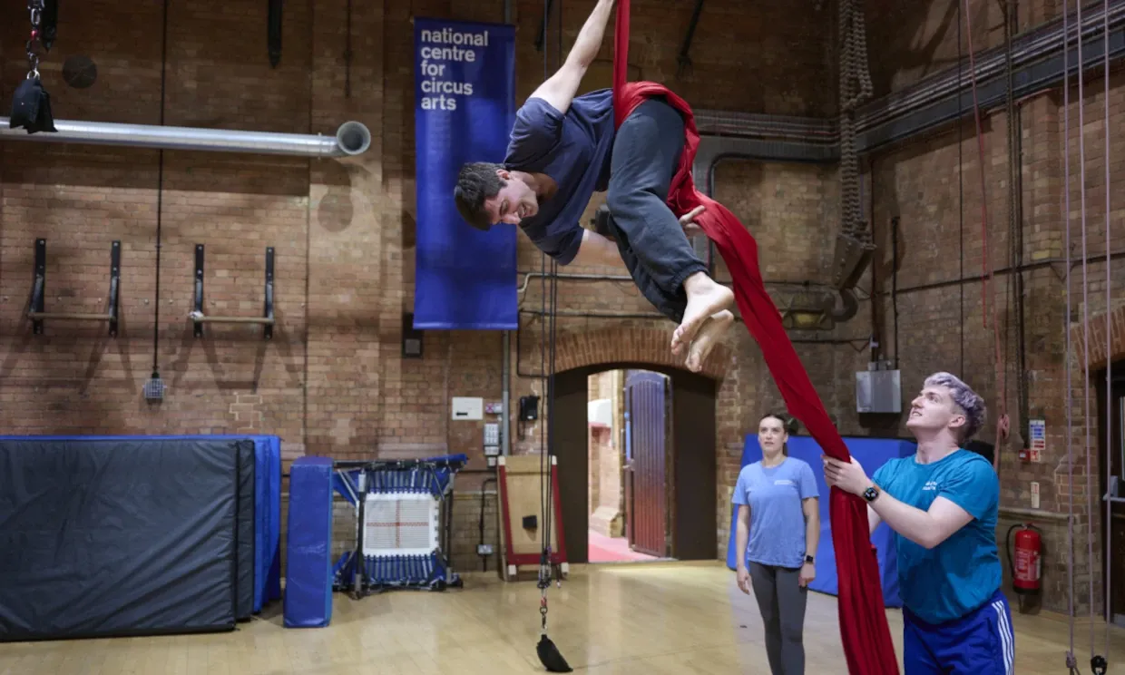 A young performer dangles from aerial ropes while being supported by someone below.